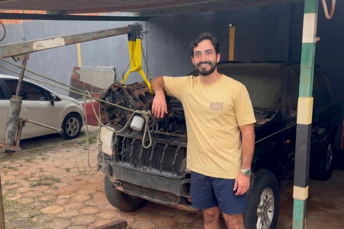 Pedro leaning on a V8 engine block hanging from a shop crane, in front of a Jeep Grand Cherokee with its front end removed